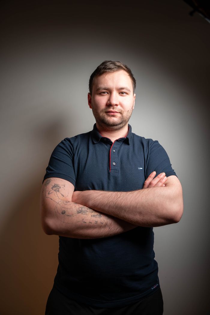 Portrait of a confident young man with tattoos standing indoors against a neutral background.